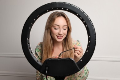 Photo of Beautiful young woman with mascara for eyelashes near white wall, view through ring lamp