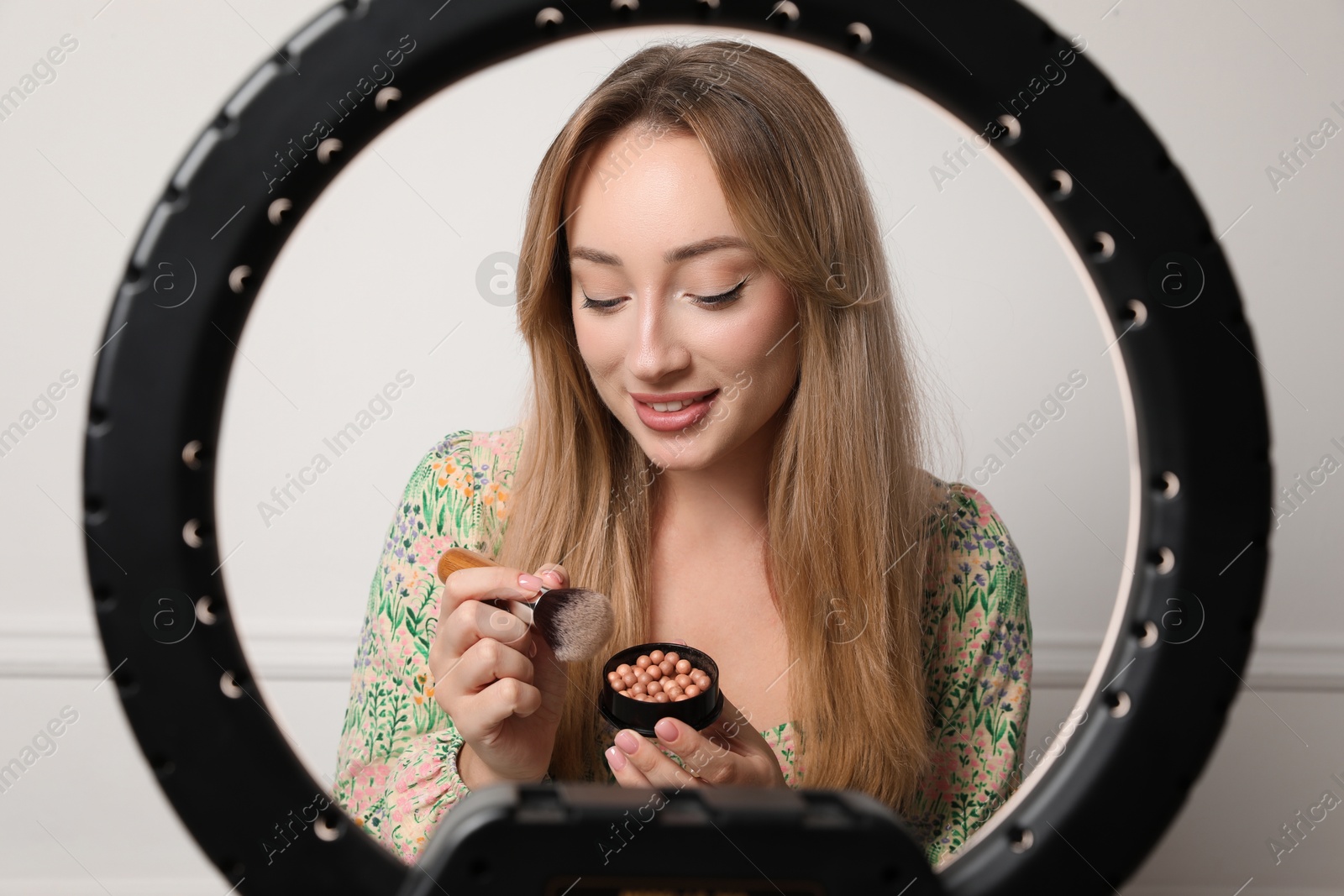 Photo of Beautiful young woman with ball blusher and brush near white wall, view through ring lamp