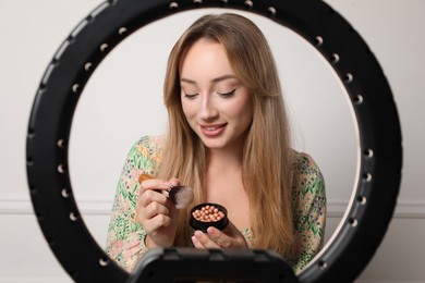 Photo of Beautiful young woman with ball blusher and brush near white wall, view through ring lamp