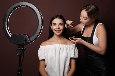 Photo of Professional makeup artist working with beautiful young woman against brown background. Using ring lamp