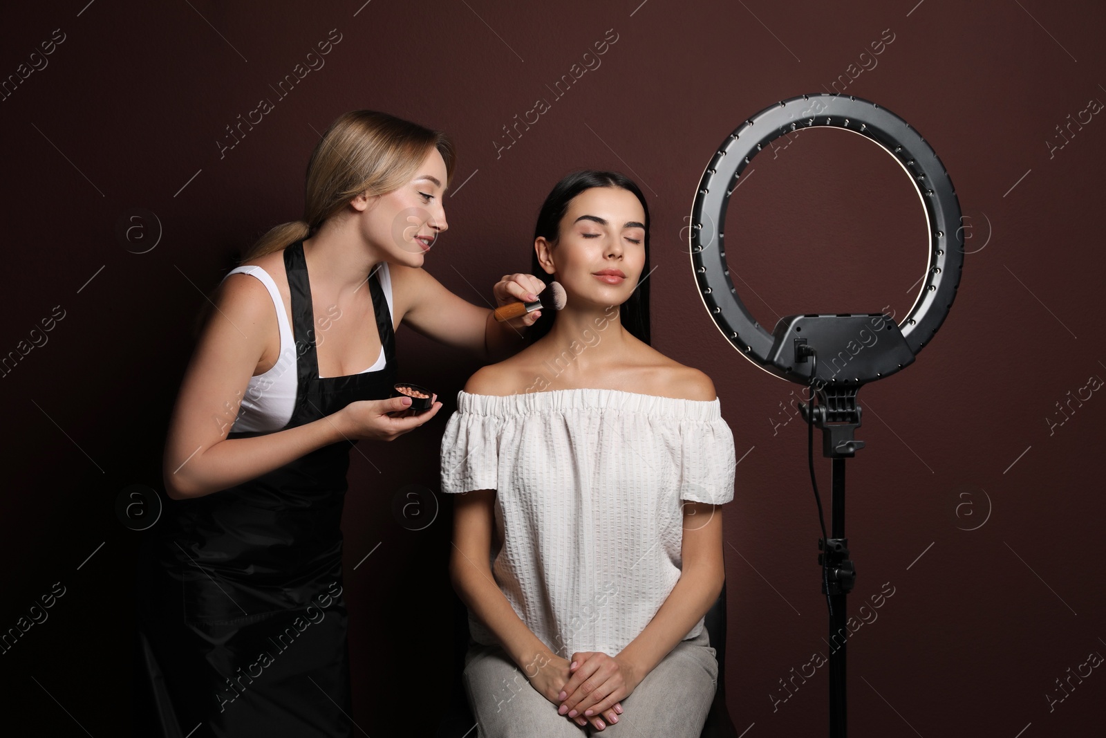 Photo of Professional makeup artist working with beautiful young woman against brown background. Using ring lamp