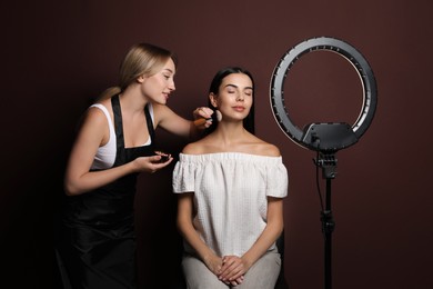 Photo of Professional makeup artist working with beautiful young woman against brown background. Using ring lamp