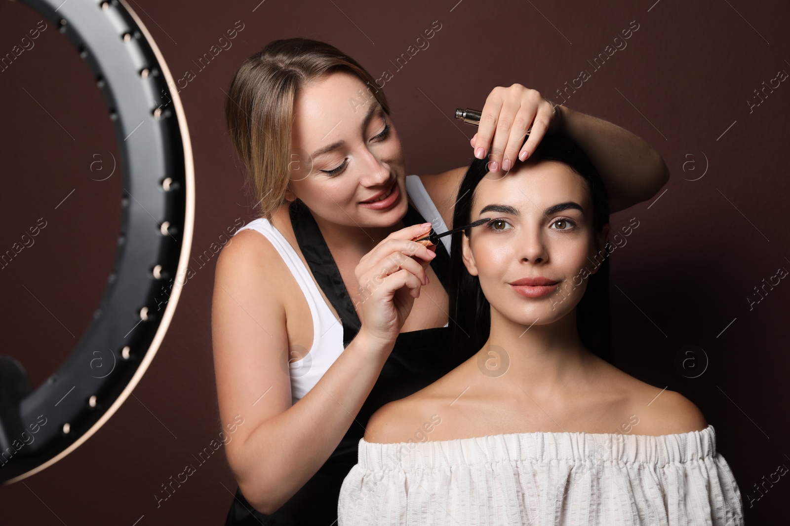 Photo of Professional makeup artist working with beautiful young woman against brown background. Using ring lamp