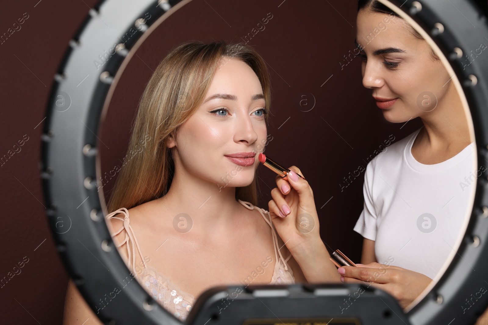 Photo of Professional makeup artist working with beautiful young woman against brown background, view through ring lamp