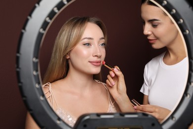 Photo of Professional makeup artist working with beautiful young woman against brown background, view through ring lamp