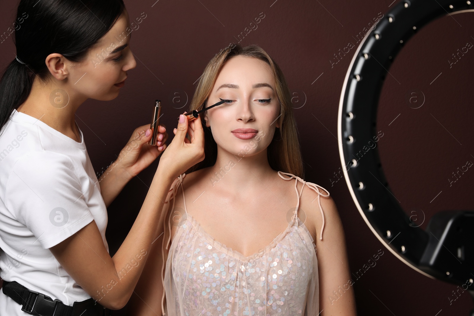 Photo of Professional makeup artist working with beautiful young woman against brown background. Using ring lamp