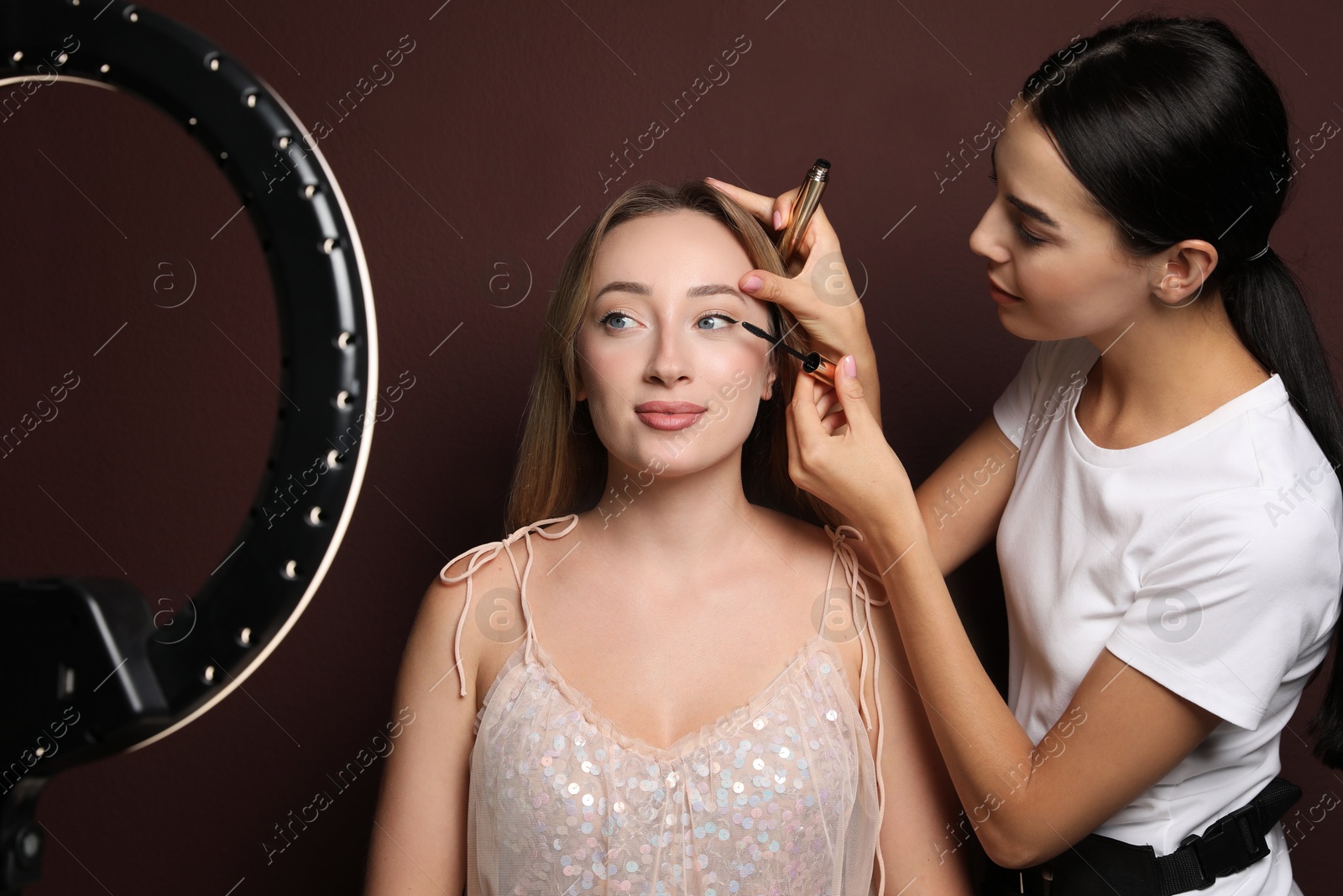 Photo of Professional makeup artist working with beautiful young woman against brown background. Using ring lamp