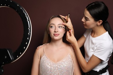 Photo of Professional makeup artist working with beautiful young woman against brown background. Using ring lamp