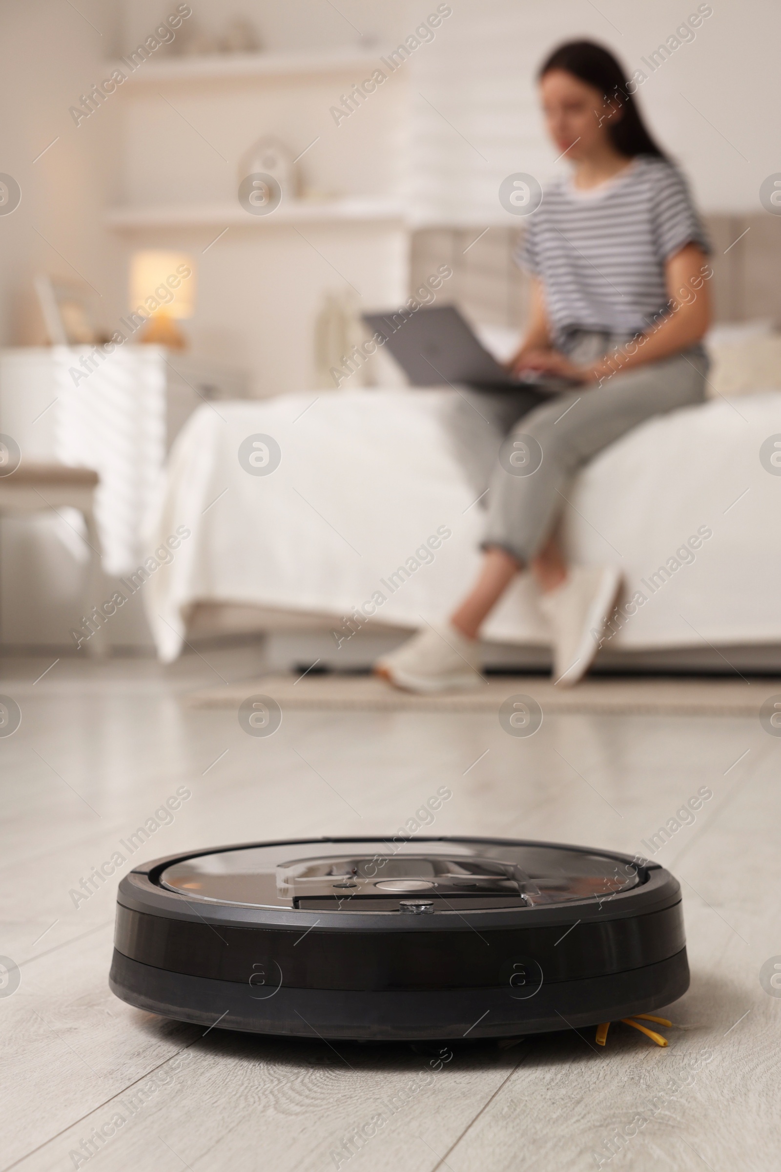 Photo of Young woman using laptop at home, focus on robotic vacuum cleaner