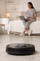 Young woman using laptop at home, focus on robotic vacuum cleaner