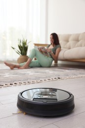 Photo of Young woman reading magazine at home, focus on robotic vacuum cleaner