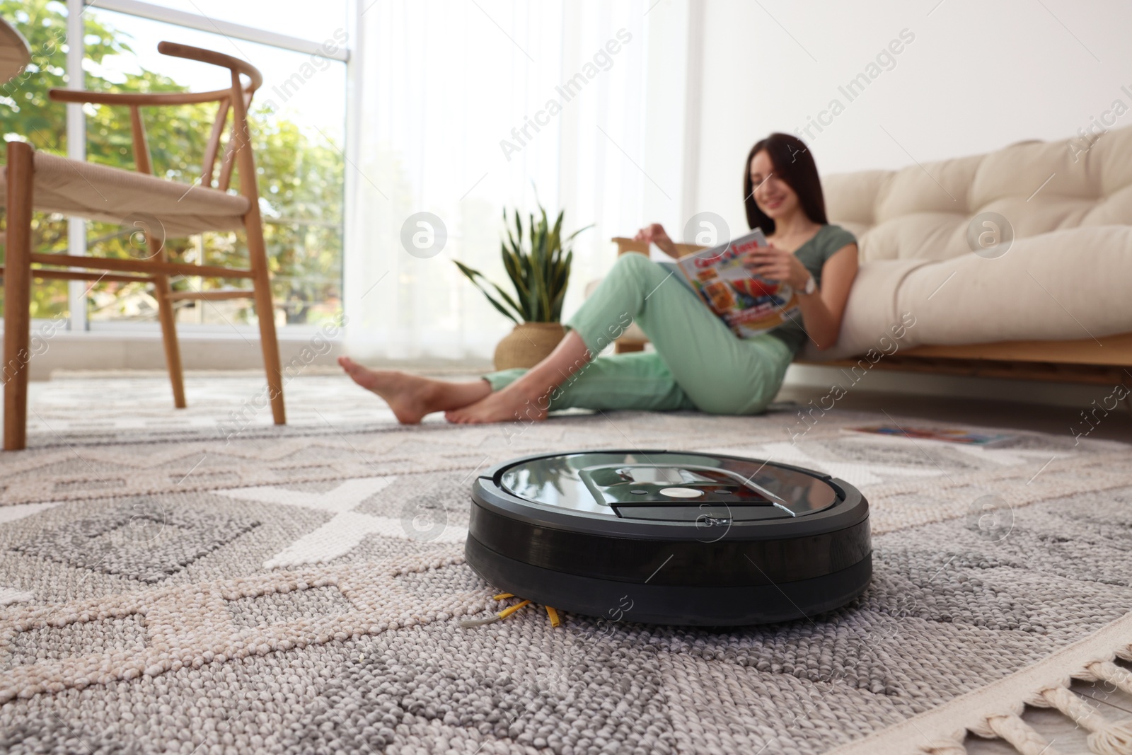 Photo of Young woman reading magazine at home, focus on robotic vacuum cleaner