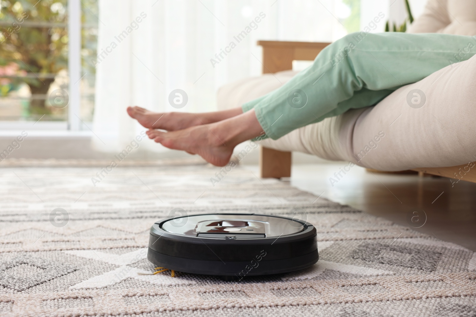 Photo of Young woman resting on sofa while robotic vacuum cleaner vacuuming rug at home, closeup