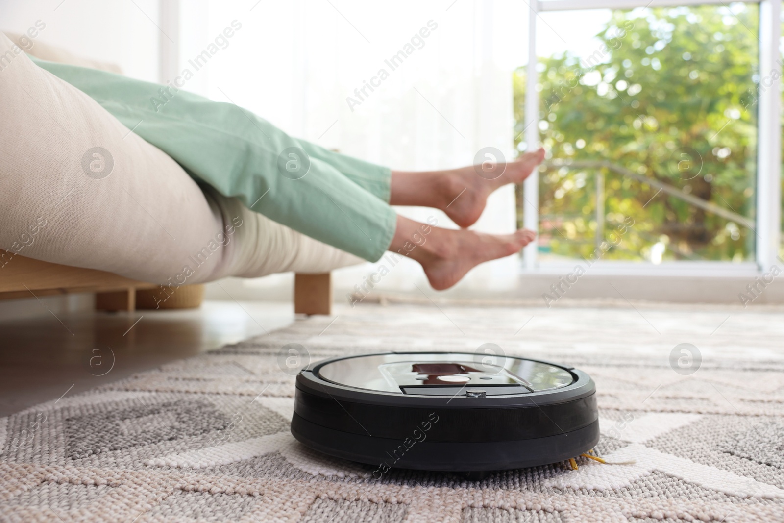 Photo of Young woman resting on sofa while robotic vacuum cleaner vacuuming rug at home, closeup