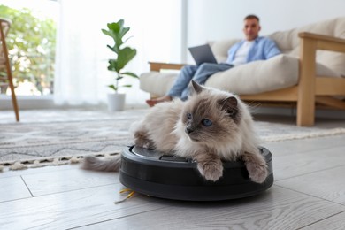 Photo of Young man using laptop at home and cute cat on robotic vacuum cleaner, selective focus