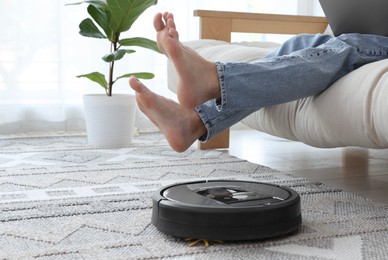 Photo of Young man using laptop while robotic vacuum cleaner vacuuming rug at home, closeup