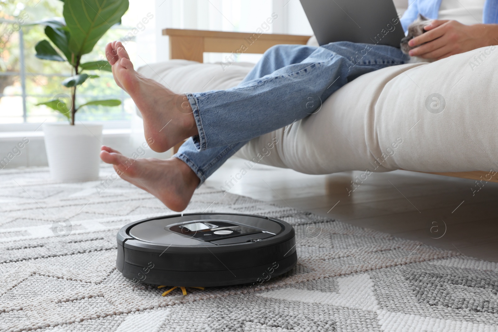 Photo of Young man using laptop while robotic vacuum cleaner vacuuming rug at home, closeup