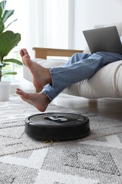 Photo of Young man using laptop while robotic vacuum cleaner vacuuming rug at home, closeup