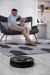 Photo of Young man reading magazine at home, focus on robotic vacuum cleaner