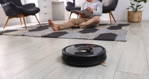 Photo of Young man reading magazine at home, focus on robotic vacuum cleaner