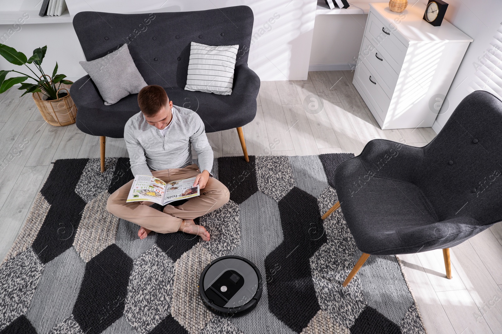 Photo of Young man reading magazine while robotic vacuum cleaner vacuuming rug at home, above view