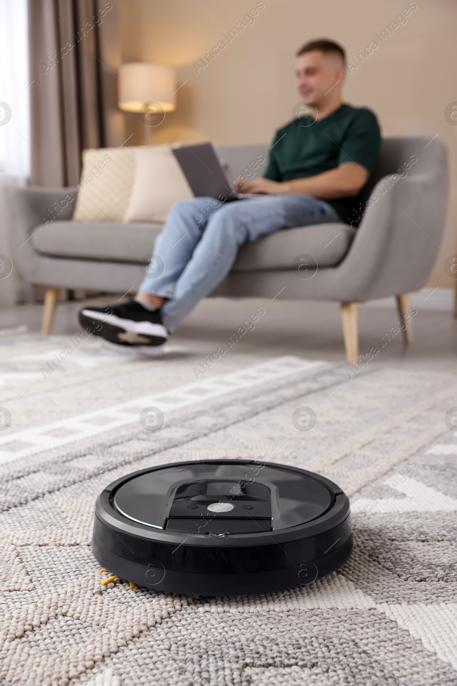 Photo of Young man using laptop at home, focus on robotic vacuum cleaner