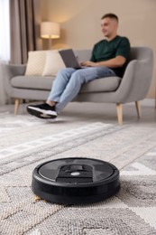 Photo of Young man using laptop at home, focus on robotic vacuum cleaner