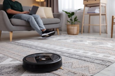 Photo of Young man using laptop at home, focus on robotic vacuum cleaner