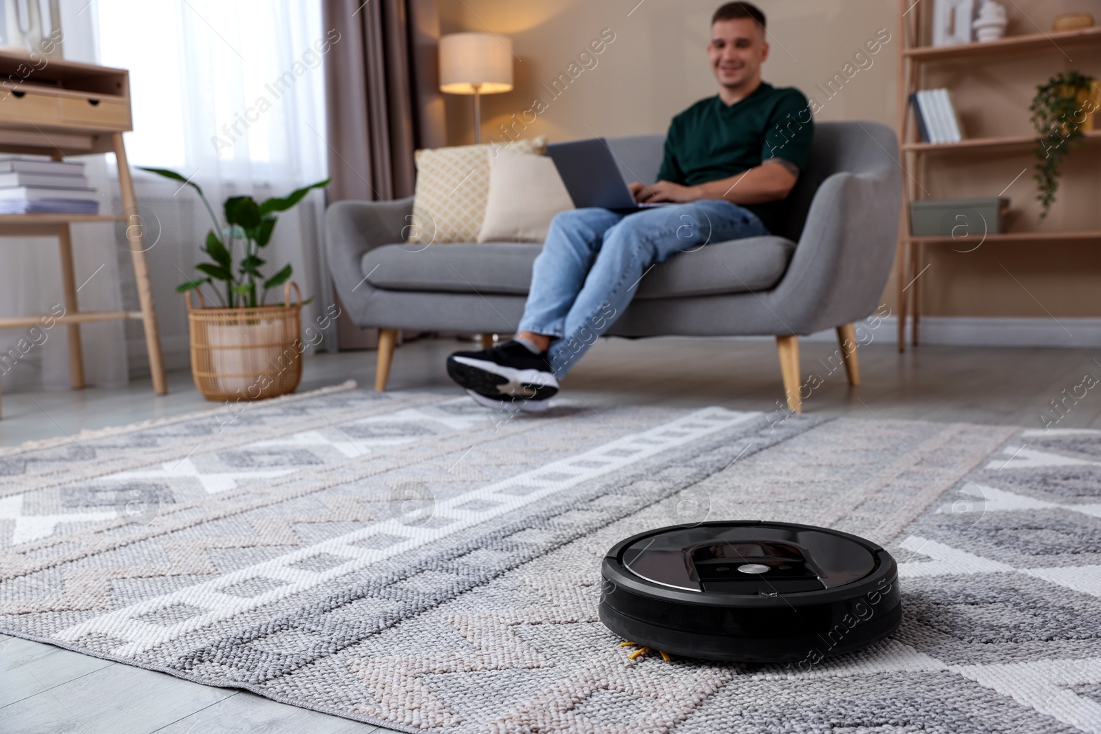 Photo of Young man using laptop at home, focus on robotic vacuum cleaner