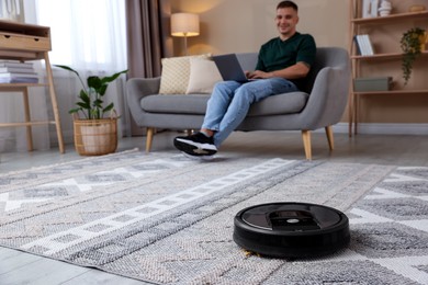 Photo of Young man using laptop at home, focus on robotic vacuum cleaner