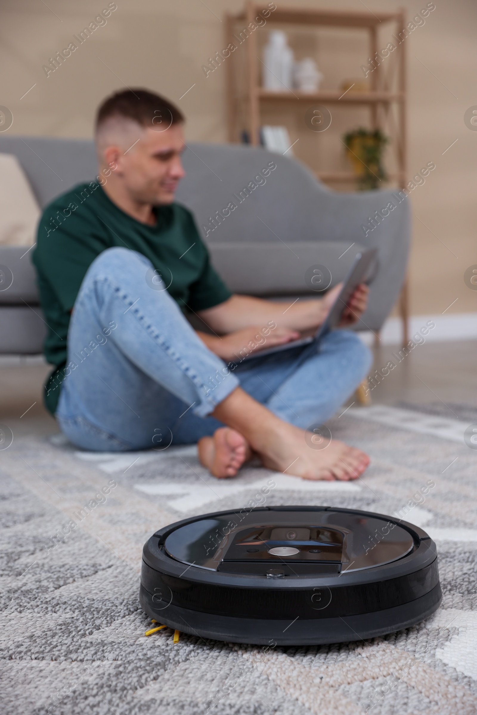 Photo of Young man using laptop at home, focus on robotic vacuum cleaner