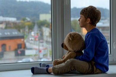 Photo of Autism concept. Lonely little boy with teddy bear near window at home