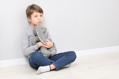 Photo of Autism concept. Lonely little boy with toy bunny on floor near grey wall at home, space for text