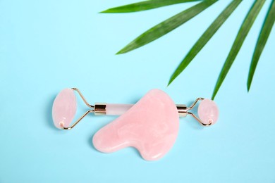 Photo of Rose quartz face roller, gua sha tool and palm leaf on light blue background