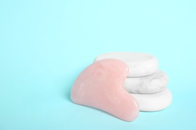 Photo of Rose quartz gua sha tool and spa stones on light blue background. Space for text