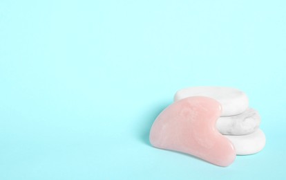 Photo of Rose quartz gua sha tool and spa stones on light blue background. Space for text