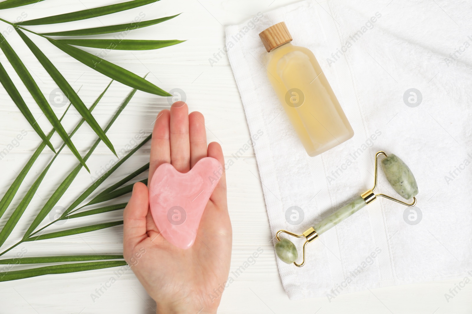 Photo of Woman with gua sha tool, face roller, cosmetic product, towel and leaves on white wooden background, top view