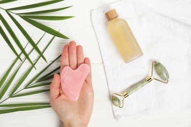 Photo of Woman with gua sha tool, face roller, cosmetic product, towel and leaves on white wooden background, top view