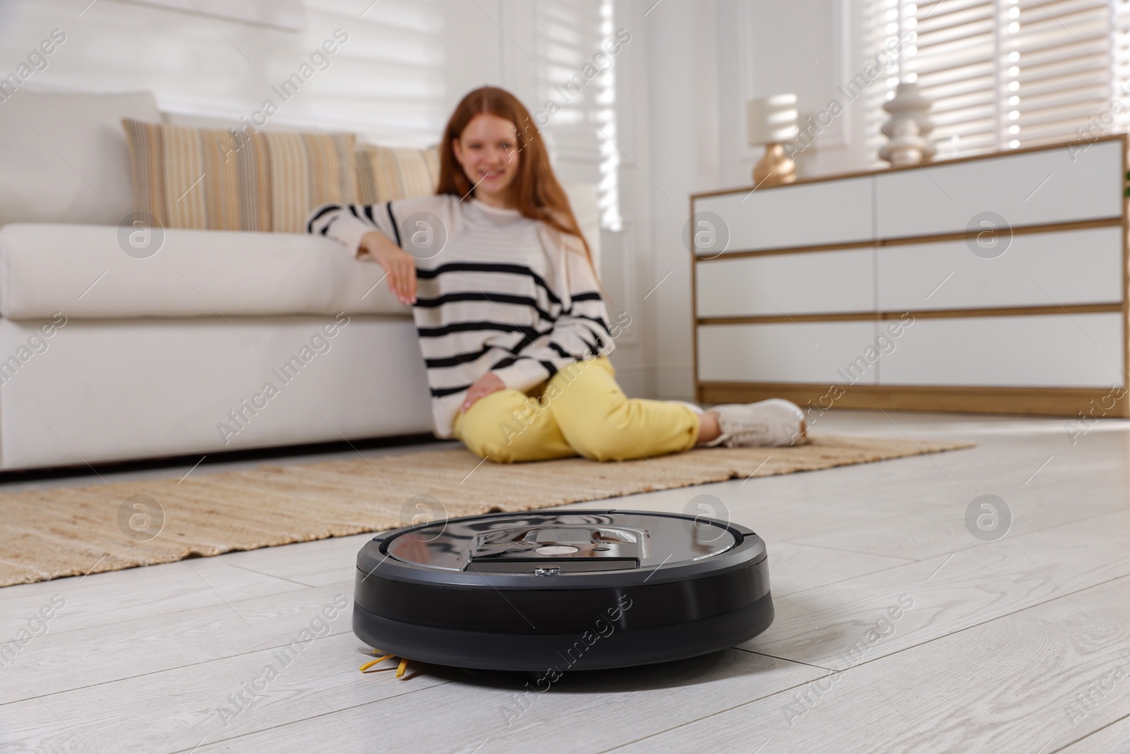 Photo of Teenage girl near sofa at home, focus on robotic vacuum cleaner