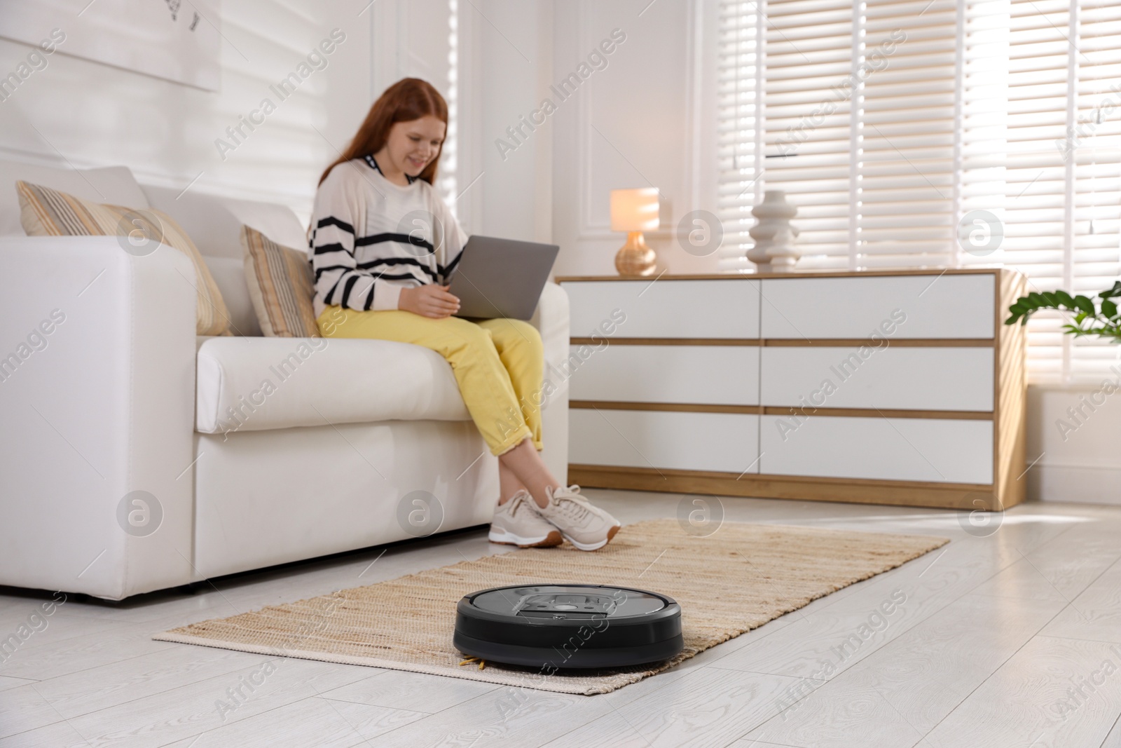 Photo of Teenage girl with laptop at home, focus on robotic vacuum cleaner