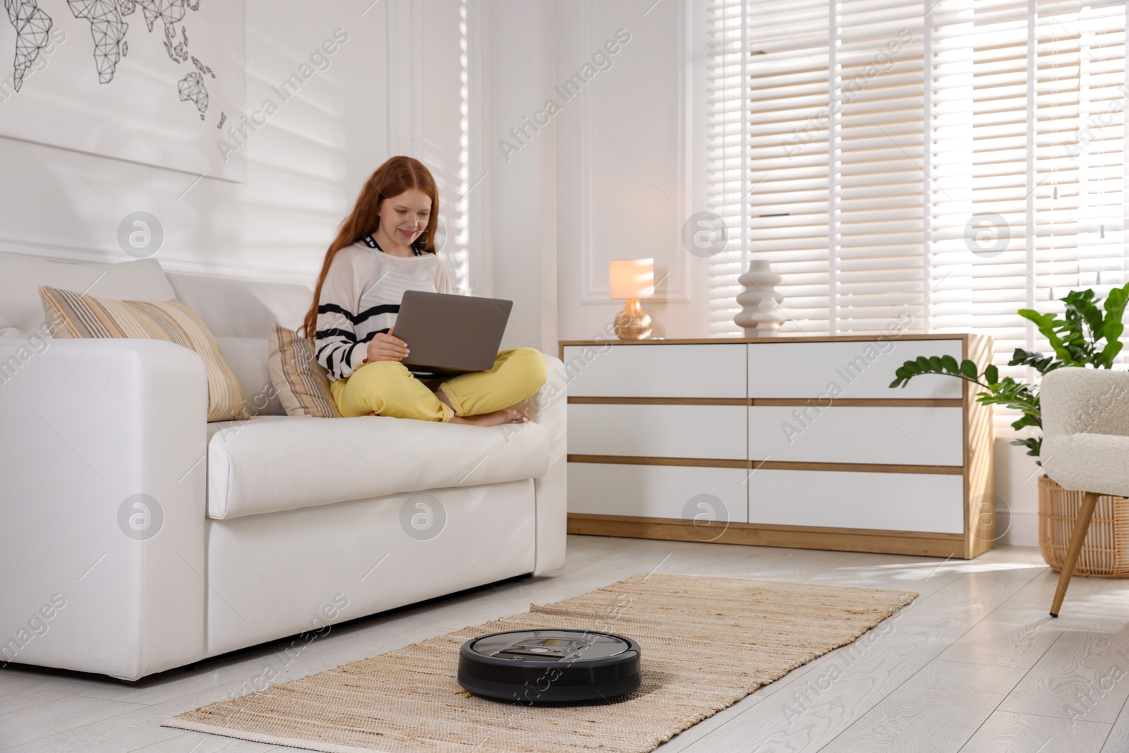 Photo of Teenage girl with laptop while robotic vacuum cleaner vacuuming rug at home