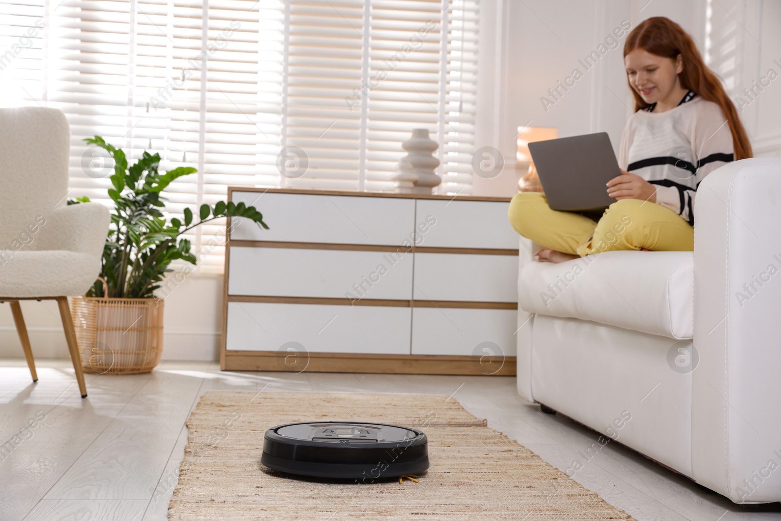 Photo of Teenage girl with laptop at home, focus on robotic vacuum cleaner