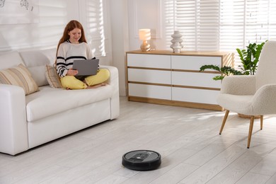 Photo of Teenage girl with laptop while robotic vacuum cleaner vacuuming floor at home