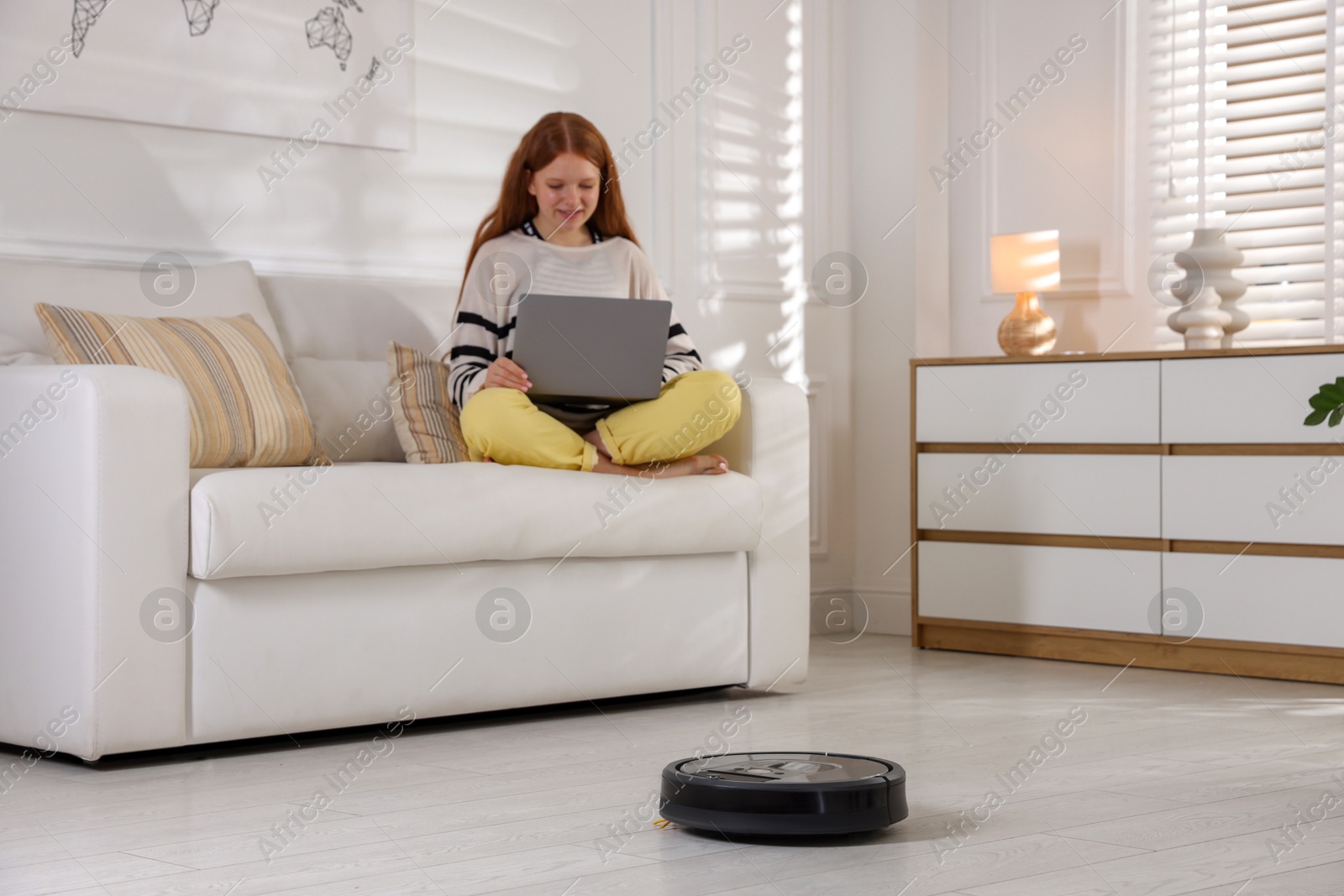 Photo of Teenage girl with laptop while robotic vacuum cleaner vacuuming floor at home