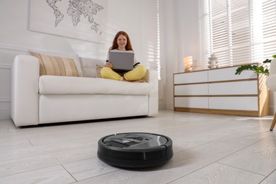 Teenage girl with laptop at home, focus on robotic vacuum cleaner