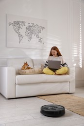 Teenage girl with laptop and her cute Chihuahua dog on sofa while robotic vacuum cleaner vacuuming floor at home