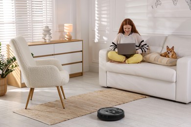 Photo of Teenage girl with laptop and her cute Chihuahua dog on sofa while robotic vacuum cleaner vacuuming floor at home