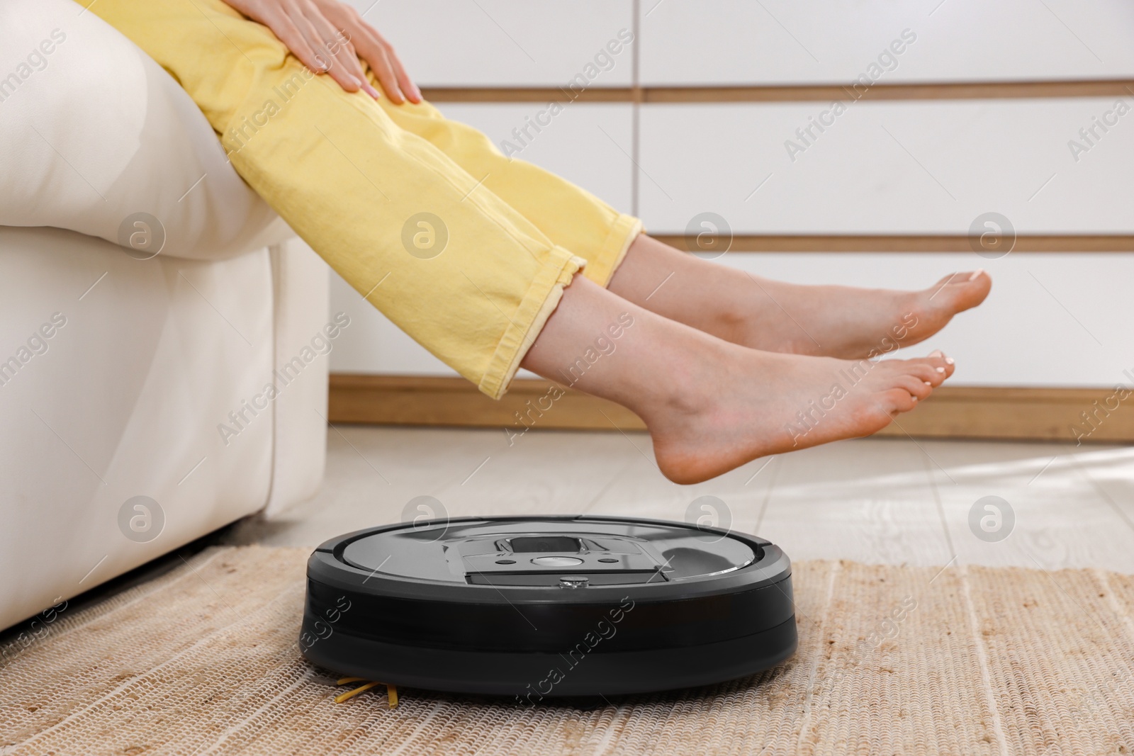 Photo of Teenage girl sitting on sofa while robotic vacuum cleaner vacuuming rug at home, closeup