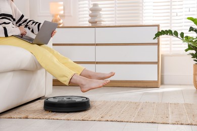 Photo of Teenage girl with laptop on sofa while robotic vacuum cleaner vacuuming rug at home, closeup