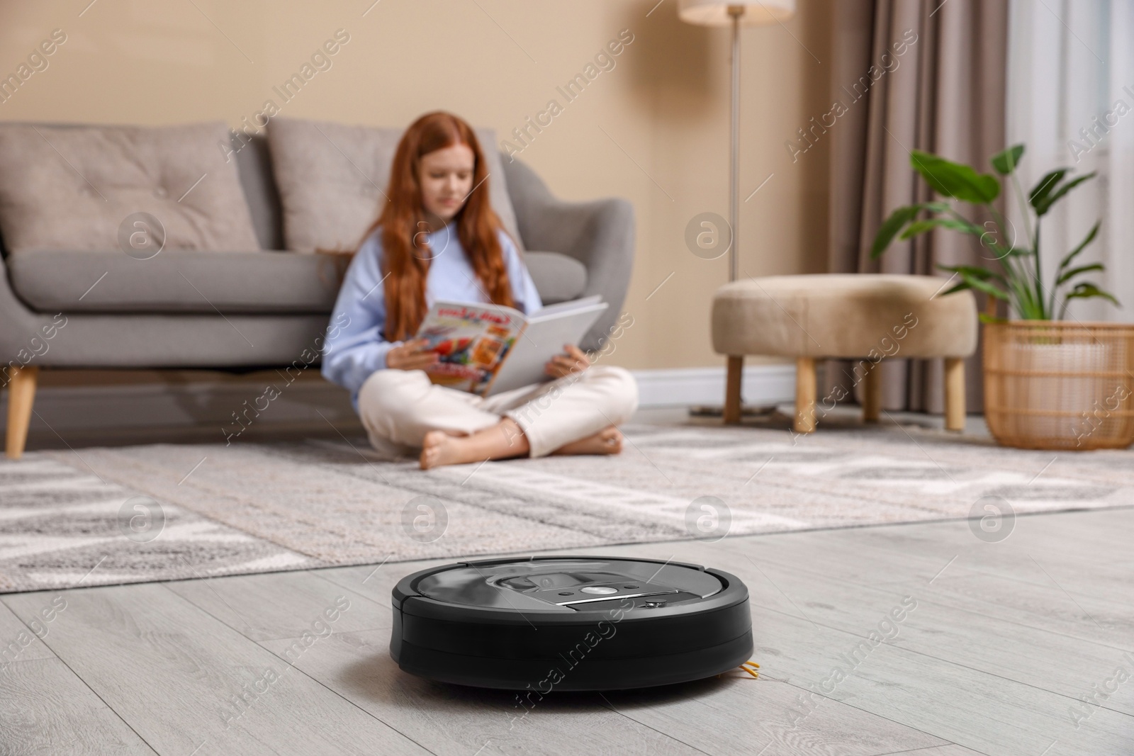 Photo of Teenage girl reading magazine while robotic vacuum cleaner vacuuming rug at home, selective focus